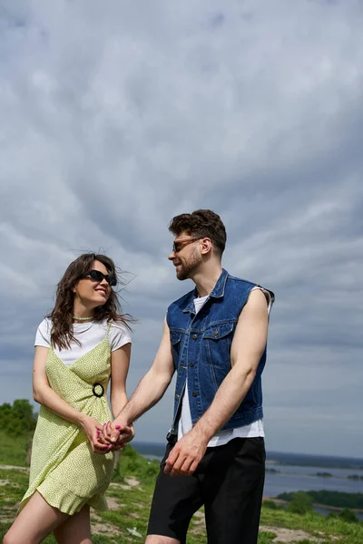 Smiling brunette woman in sunglasses and stylish sundress spending time with bearded boyfriend in summer outfit and denim vest and walking in rural setting, countryside wanderlust and love concept — Stock Photo