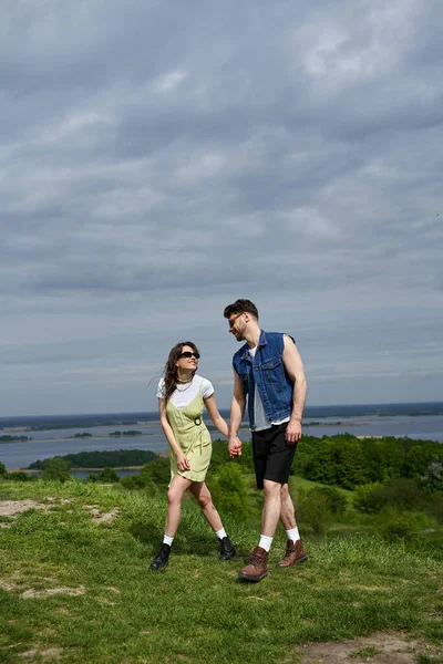 Couple romantique brune positive en lunettes de soleil et tenues d'été élégantes tenant la main et marchant ensemble sur une colline herbeuse avec vue panoramique floue en arrière-plan, promenade à la campagne — Photo de stock