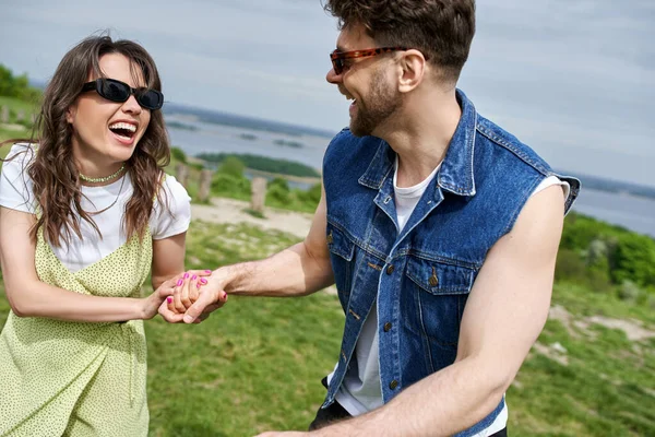 Cheerful brunette woman in trendy summer outfit and sunglasses holding hand of boyfriend in denim vest while spending time in blurred rural setting at background, countryside leisurely stroll — Stock Photo