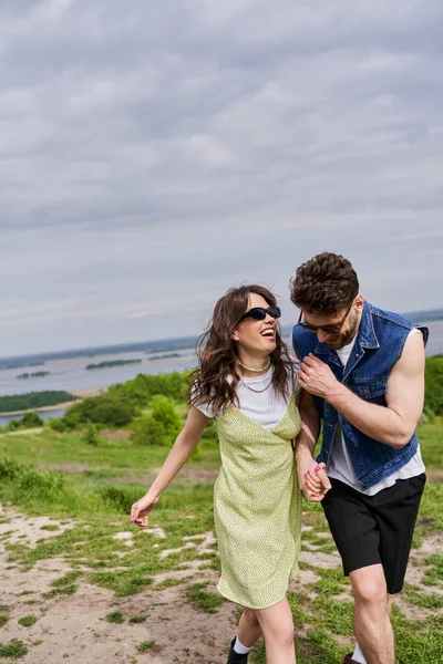 Fröhlicher brünetter Mann in stylischem Sommer-Outfit und Jeansweste hält Hand der aufgeregten Freundin in Sonnenbrille und Sonnenbrille und geht gemeinsam auf Pfad auf grasbewachsenem Hügel, Landschaft gemütlich spazieren — Stockfoto