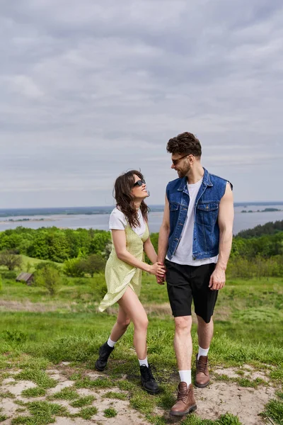 Cheerful and stylish brunette woman in sundress and boots holding hand and talking to bearded boyfriend in denim vest and sunglasses while standing in nature, countryside leisurely stroll — Stock Photo
