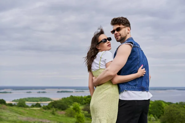 Modisches romantisches Paar in Sonnenbrille und sommerlichem Outfit, das sich umarmt und mit verschwommener ländlicher Landschaft und wolkenverhangenem Himmel im Hintergrund steht, gemütliche Spaziergänge auf dem Land, Ruhe — Stockfoto