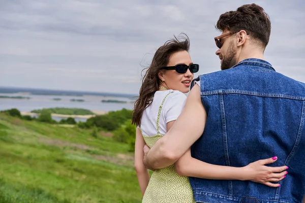 Cheerful and fashionable brunette woman in sunglasses and sundress hugging bearded boyfriend in denim vest while standing on blurred grassy hill at background, countryside retreat concept — Stock Photo