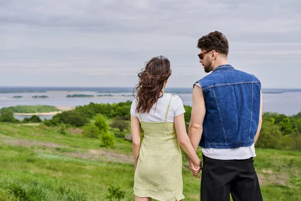Stylish bearded man in sunglasses and denim vest holding hand of brunette girlfriend in sundress and spending time together on blurred grassy hill at background, countryside retreat concept — Stock Photo