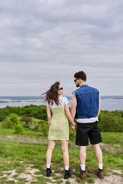 Full length of stylish brunette romantic couple in sunglasses, summer outfits and boots holding hands and spending time while standing on hill with grass, countryside retreat concept — Stock Photo