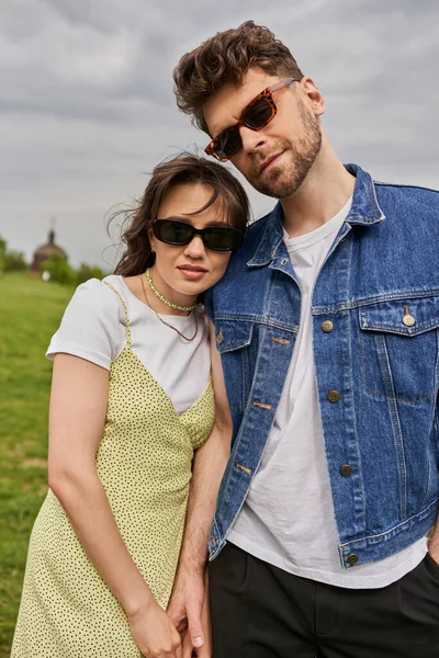 Portrait of fashionable romantic couple in sunglasses and summer outfits touching hand and posing with blurred scenic landscape at background, countryside retreat concept, tranquility — Stock Photo