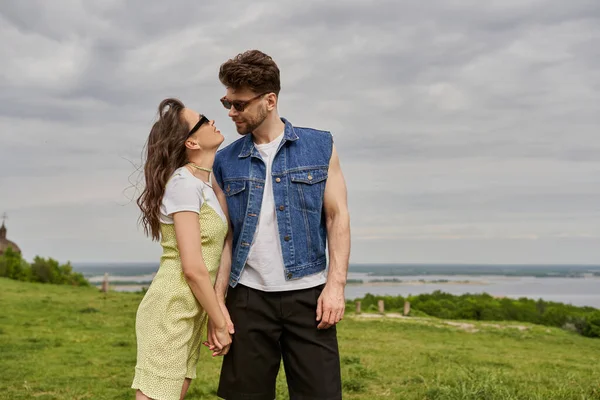 Romantic brunette woman in sunglasses and sundress holding hand of bearded boyfriend in denim vest and standing together with rural landscape and cloudy sky at background, countryside retreat concept — Stock Photo