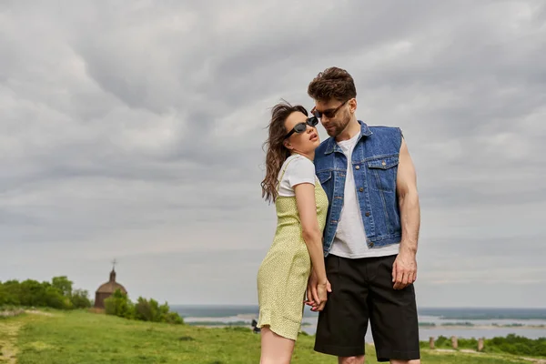 Stilvoller bärtiger Mann in Jeansweste, der die Hand einer romantischen Freundin mit Sonnenbrille und Sonnenbrille berührt, während er im Hintergrund vor ländlicher Kulisse steht, ländliches Rückzugskonzept — Stockfoto