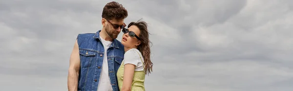 Trendy bearded man in sunglasses and denim vest looking at brunette girlfriend in stylish sundress and standing with blurred cloudy sky at background, countryside retreat concept, banner — Stock Photo