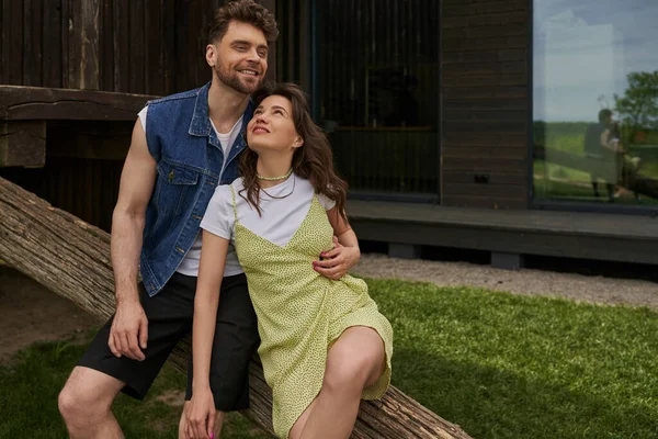 Positive an bearded man in denim vest hugging stylish girlfriend in sundress and sitting together on wooden log while relaxing in rural setting, countryside retreat concept — Stock Photo
