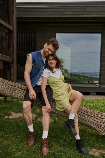 Full length of stylish and fashionable romantic couple in summer outfits and boots resting on wooden log with house at background in rural setting, outdoor enjoyment concept, tranquility — Stock Photo