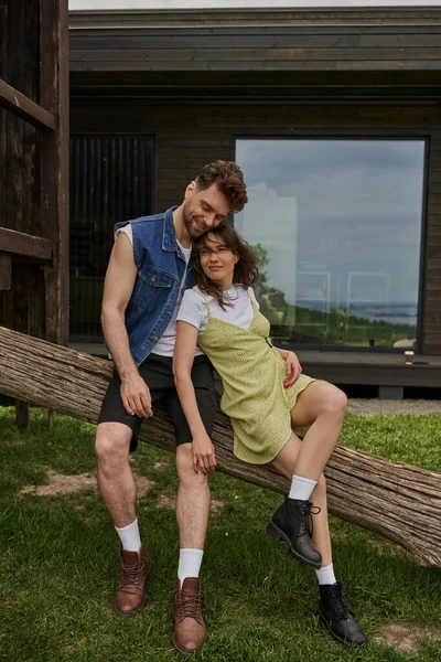 Longitud completa de pareja romántica elegante y de moda en trajes de verano y botas que descansan en el registro de madera con la casa en el fondo en el entorno rural, concepto de disfrute al aire libre, tranquilidad - foto de stock