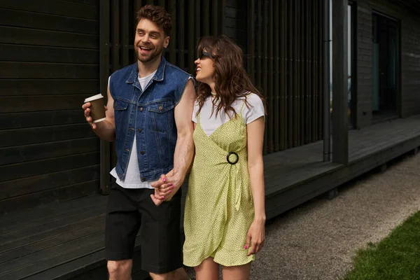 Smiling and stylish bearded man holding coffee to go and hand of brunette girlfriend in sunglasses and summer dress while walking near house in rural setting, outdoor enjoyment concept, tranquility — Stock Photo