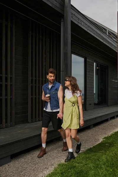 Homme élégant en tenue d'été tenant café pour aller et main de petite amie gaie dans des lunettes de soleil et robe de soleil tout en marchant près de la maison en bois dans un cadre rural, concept de plaisir en plein air, tranquillité — Photo de stock