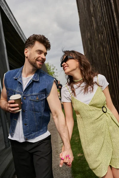 Весела романтична пара в стильному літньому вбранні тримає руки і каву, щоб піти під час розмови і прогулянки між дерев'яними будинками в сільській місцевості на фоні, концепція насолоди на відкритому повітрі — стокове фото