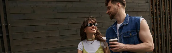 Femme brune souriante en lunettes de soleil et tenue d'été regardant petit ami barbu tenant du café à emporter tout en marchant près de la maison en bois à l'arrière-plan, concept de plaisir en plein air, bannière — Photo de stock