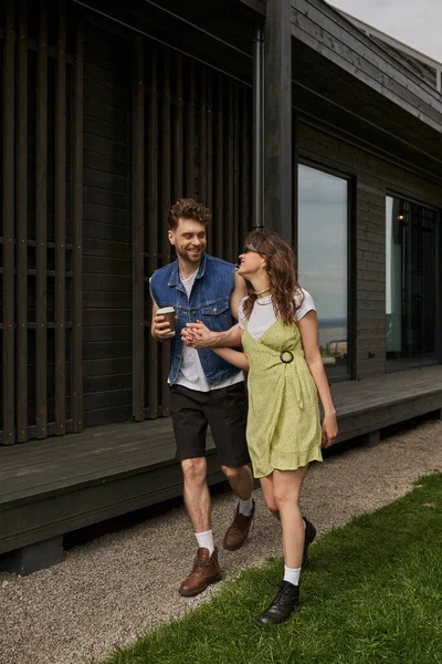 Trendy and stylish couple in summer outfits and boots holding takeaway coffee and talking while walking together near wooden house in rural setting, outdoor enjoyment concept — Stock Photo
