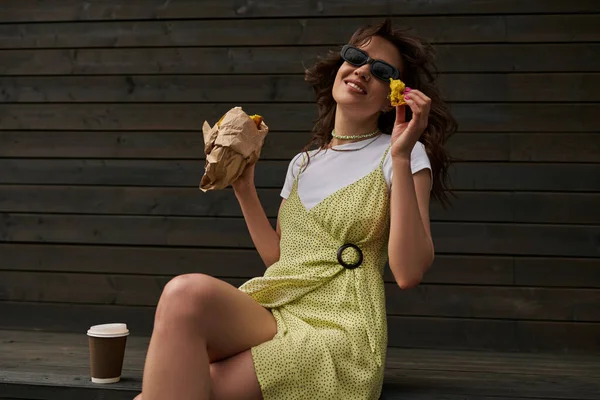 Cheerful and stylish brunette woman in sunglasses and sundress holding bun in paper bag while sitting near coffee to go and wooden house in rural setting, summer vibes concept — Stock Photo