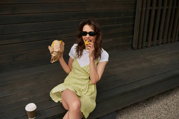 Stylish brunette woman in sundress and sunglasses eating fresh bun while sitting near takeaway coffee while relaxing near wooden house in rural setting, summer vibes concept — Stock Photo