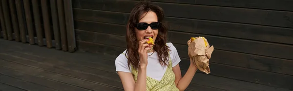 Femme brune branchée en lunettes de soleil et robe de soleil élégante mangeant un pain frais savoureux tout en étant assise près d'une maison en bois dans un cadre rural, concept d'ambiance estivale, bannière, tranquillité — Photo de stock