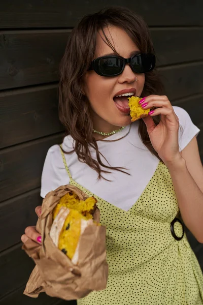Portrait of stylish brunette woman in sunglasses in summer outfit eating fresh bun in craft package while standing near wooden house in rural setting, summer vibes concept, tranquility — Stock Photo