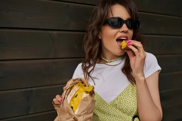 Portrait de femme brune en tenue d'été et lunettes de soleil mangeant un pain frais savoureux tout en se tenant près de la maison floue dans un cadre rural, concept d'ambiance estivale, tranquillité — Photo de stock