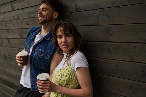Stylish brunette woman in summer outfit holding coffee to go and looking away near cheerful bearded boyfriend in denim vest and wooden house at background, carefree moments concept, tranquility — Stock Photo