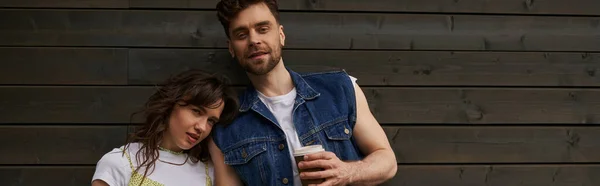Smiling bearded and stylish man in denim vest holding coffee to go and looking at camera near stylish girlfriend and wooden house in rural setting, carefree moments concept, banner — Stock Photo