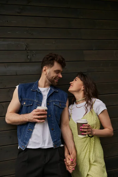 Cheerful and stylish romantic couple in summer outfits looking at each other and holding coffee to go while standing near wooden house in rural setting, carefree moments concept — Stock Photo