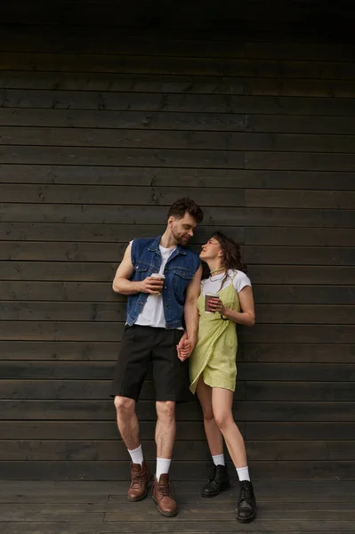 Повна довжина модної романтичної пари в чоботях і літньому вбранні, що тримає каву, щоб йти і дивитися один на одного, стоячи поруч з дерев'яним будинком, безтурботні моменти концепції — стокове фото