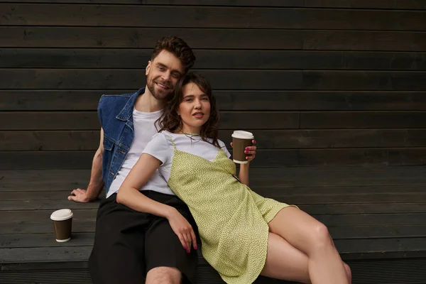Fashionable stylish and romantic couple in summer outfits smiling at camera while holding coffee to go and sitting near wooden house in rural setting, carefree moments concept, tranquility — Stock Photo