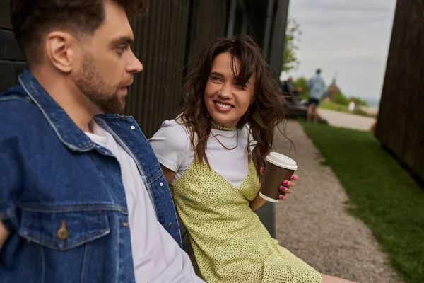 Positive brunette woman in stylish summer outfit holding coffee to go and looking at blurred bearded boyfriend in denim vest near wooden house in rural setting, carefree moments concept — Stock Photo