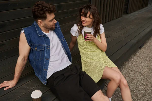 Homme barbu souriant et élégant en gilet en jean parlant à une petite amie brune en robe de soleil tenant du café pour aller s'asseoir près d'une maison en bois dans un cadre rural, concept de moments insouciants, tranquillité — Photo de stock