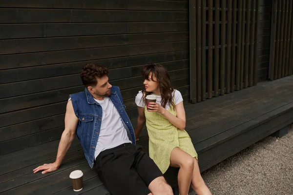 Stylish brunette woman in sundress holding takeaway coffee and looking at bearded boyfriend in denim vest while sitting near wooden house in rural setting, carefree moments concept — Stock Photo