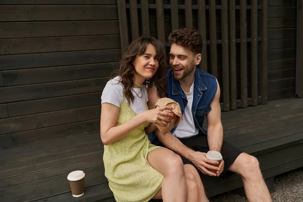 Cheerful brunette woman in sundress holding craft package and looking at camera near bearded boyfriend with coffee to go and wooden house in rural setting, serene ambiance concept — Stock Photo