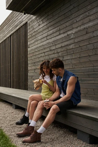 Smiling brunette woman in summer outfit and boots holding fresh bun in craft package and sitting near stylish boyfriend with coffee to go near wooden house, serene ambiance concept — Stock Photo