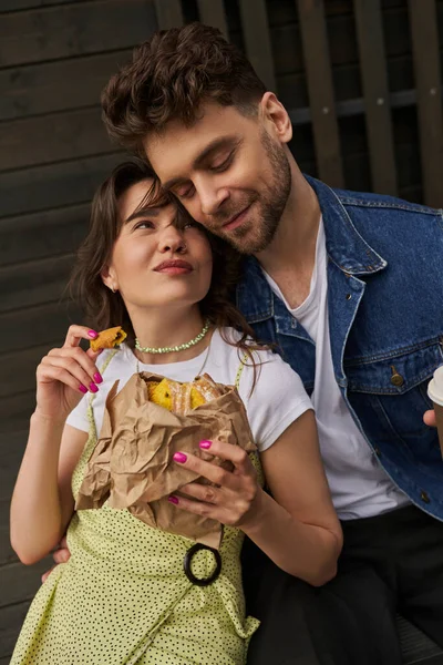 Portrait of joyful brunette woman in sundress holding fresh bun in craft package and looking at stylish boyfriend in denim vest while sitting near wooden house at background, serene ambiance concept — Stock Photo