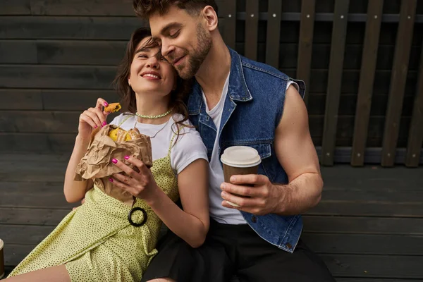 Радостная и стильная женщина в сарафане, держа в руках свежую булочку и сидя рядом бородатый бойфренд в джинсовой жилетке с кофе на вынос и деревянный дом на заднем плане, спокойная концепция атмосферы — стоковое фото