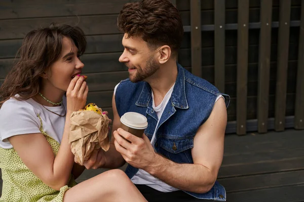 Улыбающийся бородатый мужчина в джинсовой жилетке держит кофе, чтобы пойти и поговорить с подругой в летнем наряде едят свежую булочку и сидят рядом с деревянным домом на заднем плане, спокойная концепция атмосферы — стоковое фото