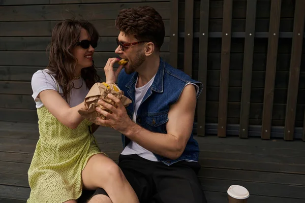 Positive brunette woman in sunglasses and sundress feeding boyfriend with tasty bun and sitting near coffee to go and wooden house at background, serene ambiance concept, tranquility — Stock Photo