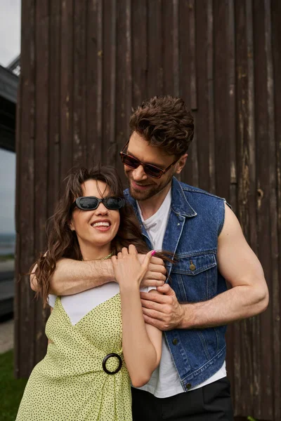 Positive brunette man in sunglasses and denim vest embracing girlfriend in stylish sundress and standing near wooden house at background in rural setting, countryside exploration concept — Stock Photo