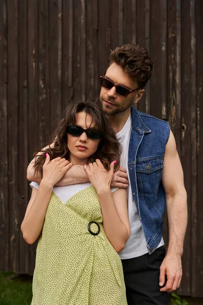 Portrait de l'homme brune élégant en tenue d'été étreignant petite amie en robe de soleil et lunettes de soleil tout en se tenant ensemble près de la maison en bois à l'arrière-plan, concept d'exploration de la campagne — Photo de stock