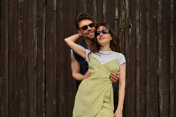 Joyful brunette man in sunglasses hugging trendy girlfriend in sundress and spending time together near wooden house in rural setting, countryside exploration concept, tranquility — Stock Photo