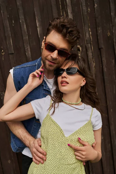 Portrait of stylish bearded man in sunglasses and denim vest embracing brunette girlfriend in sundress and standing together near wooden building outdoors, countryside exploration concept — Stock Photo