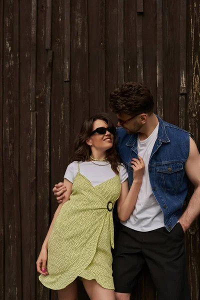 Homme barbu à la mode en lunettes de soleil et gilet en denim embrassant petite amie joyeuse en robe de soleil et debout ensemble près de la maison en bois à l'arrière-plan, concept d'exploration de la campagne — Photo de stock