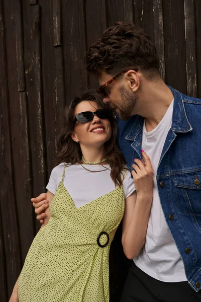 Positive brunette woman in stylish sundress and sunglasses touching bearded boyfriend in denim vest while spending time near wooden house at background, countryside exploration concept — Stock Photo