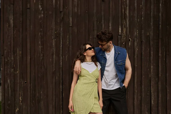 Модный брюнет в солнцезащитных очках и джинсовом жилете обнимает улыбающуюся подругу в раздевалке и держит руку в кармане, стоя рядом с деревянным домом, загородной концепцией — стоковое фото