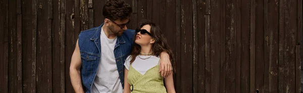 Brunette man in sunglasses and denim vest hugging joyful girlfriend in sundress and standing together near wooden house at background, countryside exploration concept, banner — Stock Photo