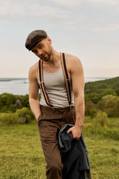 Portrait of fashionable and bearded man in newsboy cap and vintage outfit holding hand in pocket and jacket while standing with rural landscape at background, scenic countryside concept — Stock Photo