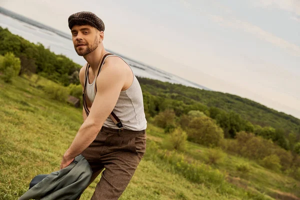 Trendy bearded man in newsboy cap and suspenders holding jacket and looking at camera while posing in vintage outfit with landscape at background, scenic countryside concept — Stock Photo